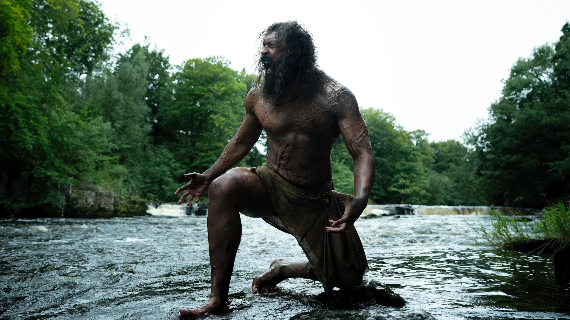 Samson, played by Chi Lewis-Parry, kneeling on a rock in a fast river and roaring, trees on both banks, in 28 Years Later: The Bone Temple