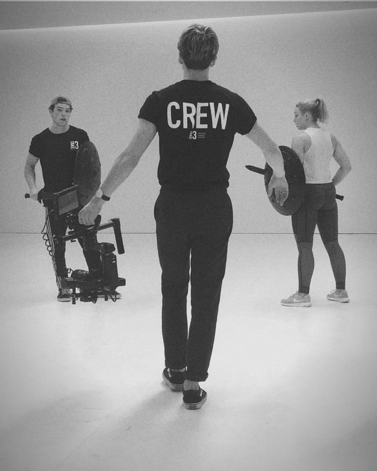 Three of the Take 3 team in a white studio: two in black Crew t-shirts carrying a camera rig and a reflector, one in workout gear