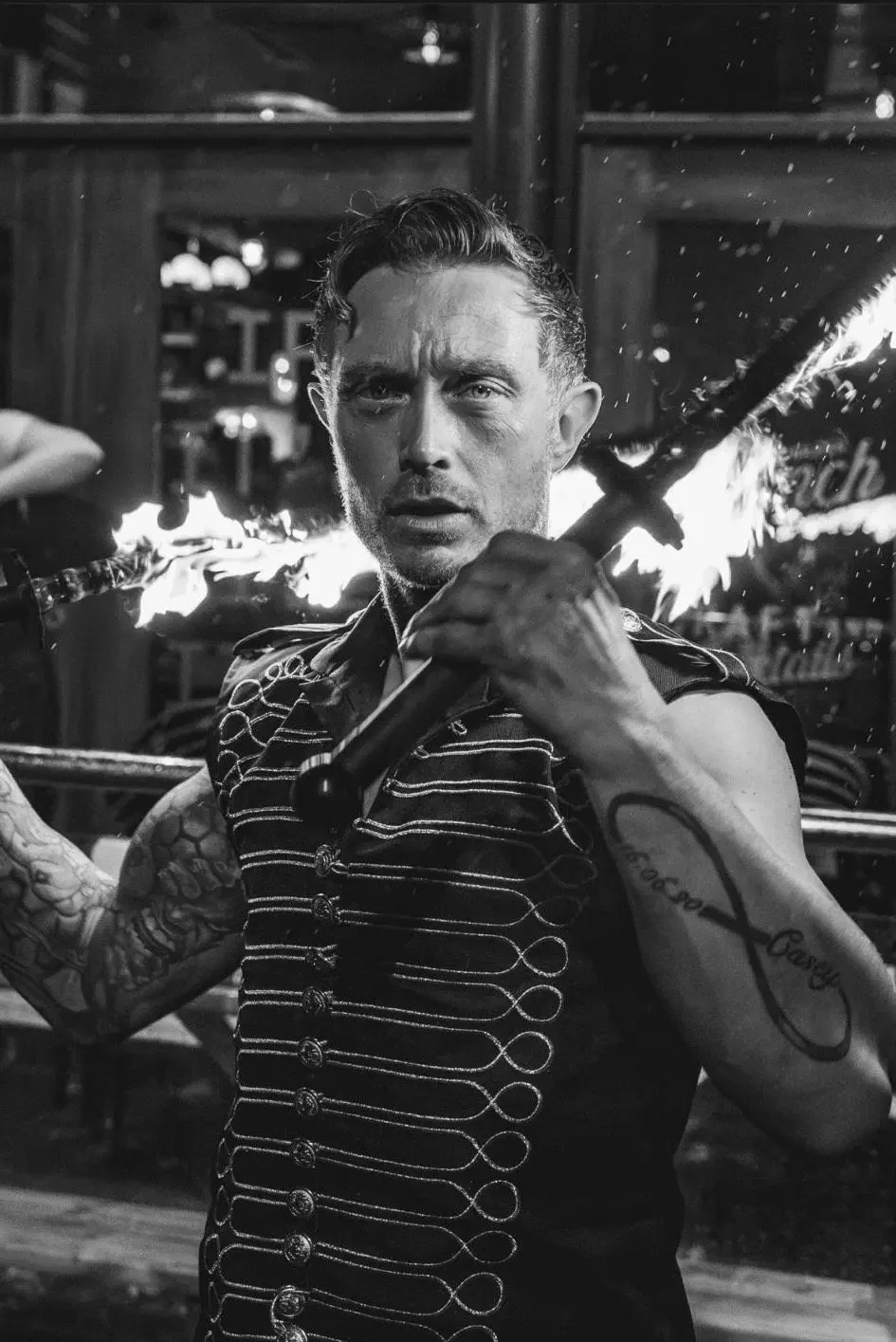 A fire performer, photographed in black and white, in an embroidered waistcoat holding a lit fire staff across his shoulders, sparks in the air behind him.