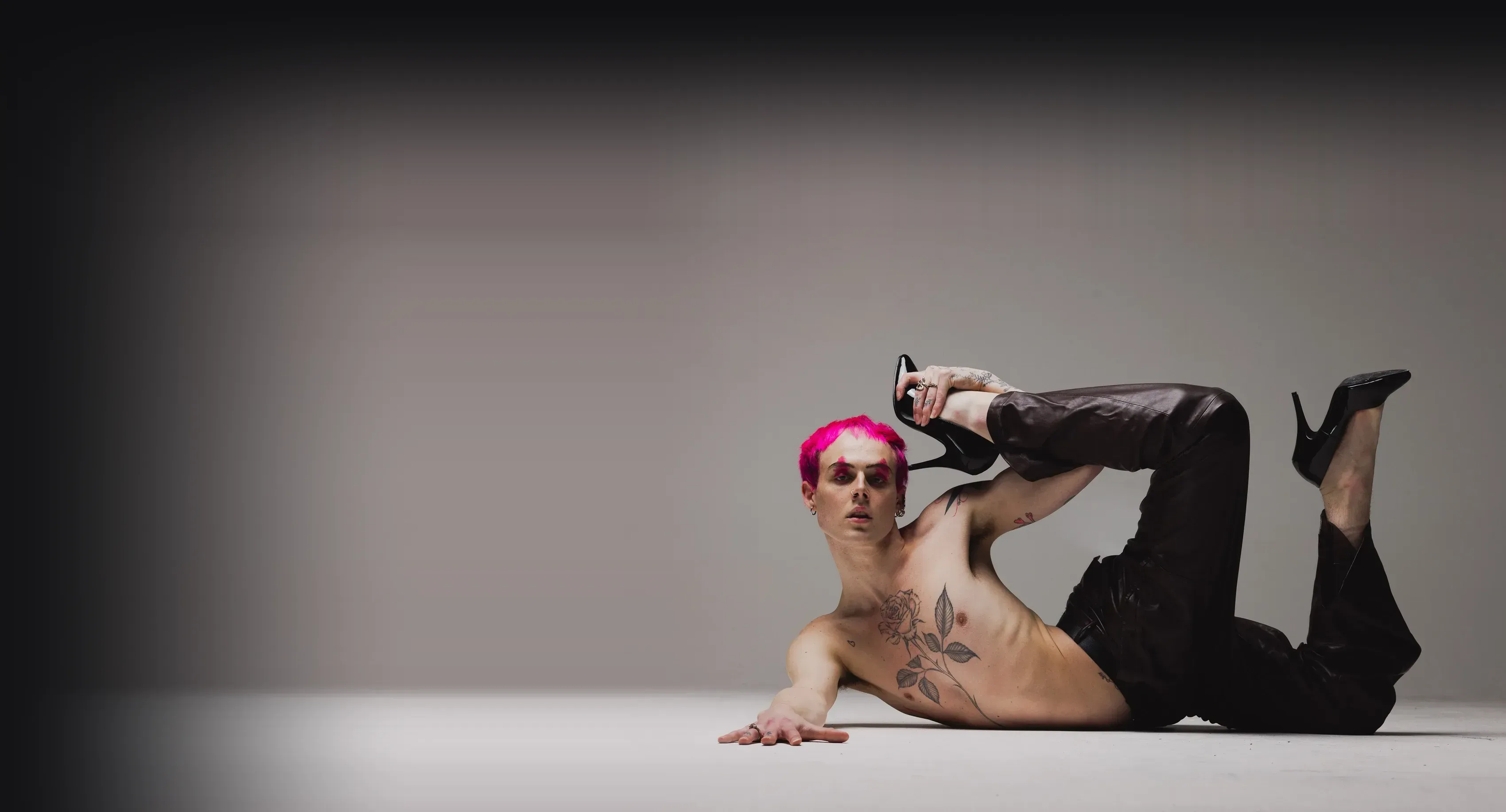A performer with vivid pink hair and tattoos lying on a studio floor in leather trousers, one leg raised and a patent heel held in hand.