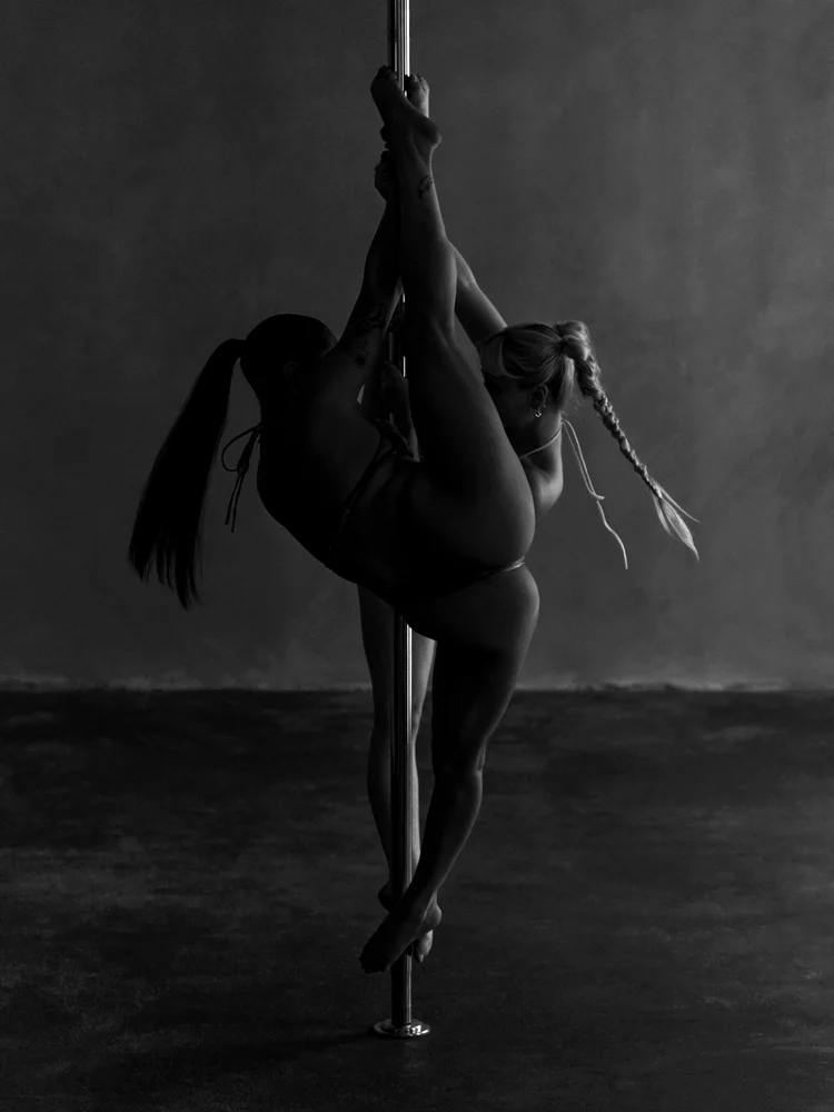 Two pole artists mirrored on a single pole in a low-lit studio, photographed in black and white, legs extended in a split.