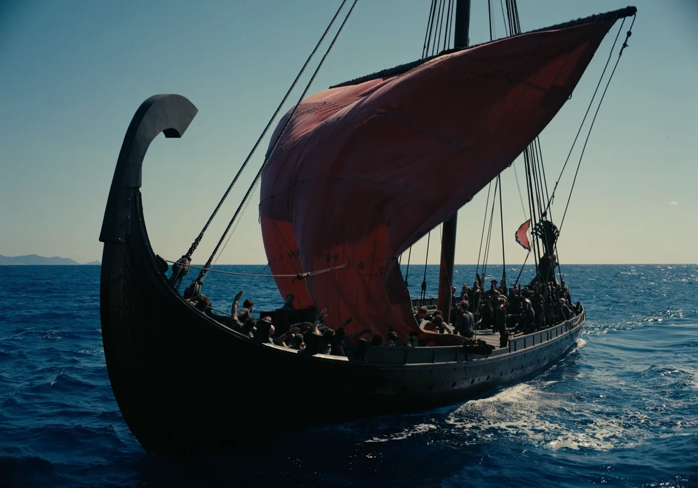 A longship under a full red sail crossing open blue sea, its crew crowded along the deck