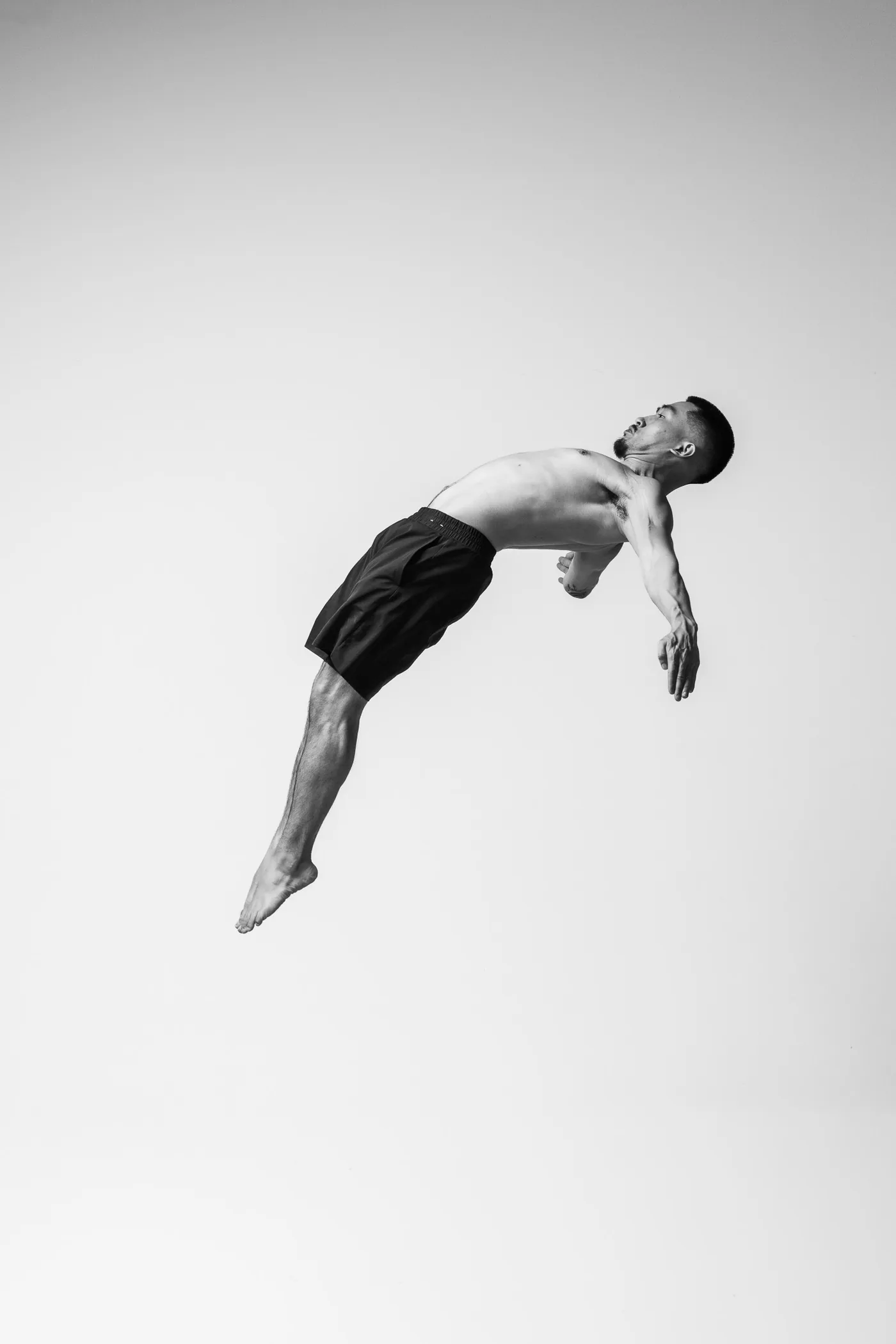 An acrobat mid-air in a back layout, arms swept behind him and feet pointed, against a plain studio wall in black and white.