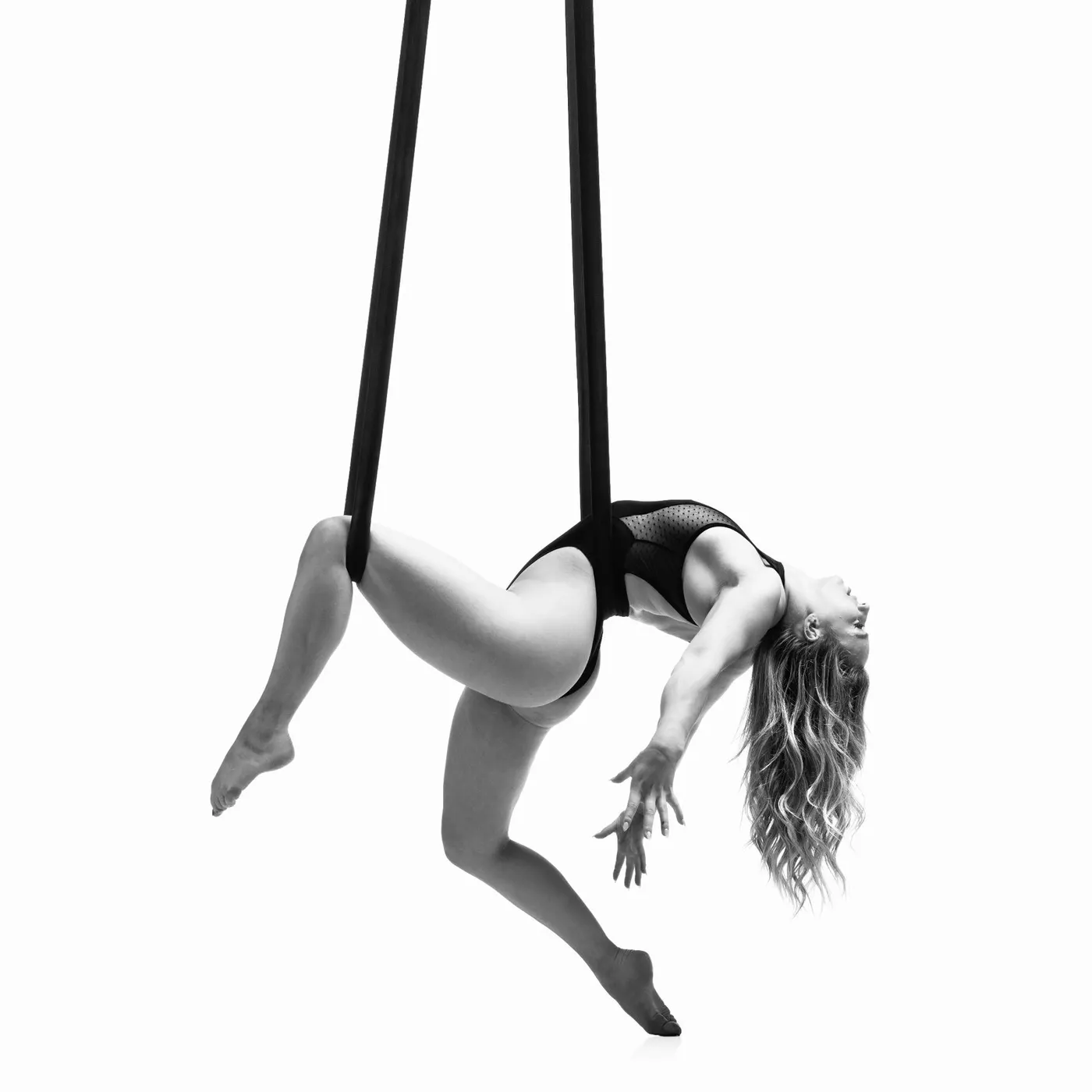 An aerialist hanging from two straps in a deep backbend, hair falling towards the floor, photographed in black and white against white.