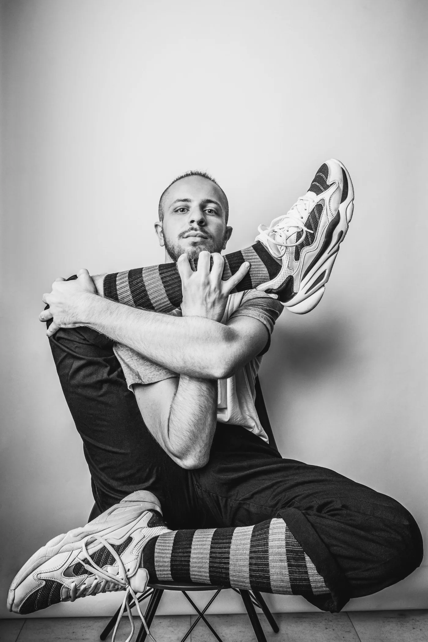 A contortionist seated on a chair with one leg folded up behind his shoulder, looking straight at the camera, in black and white.
