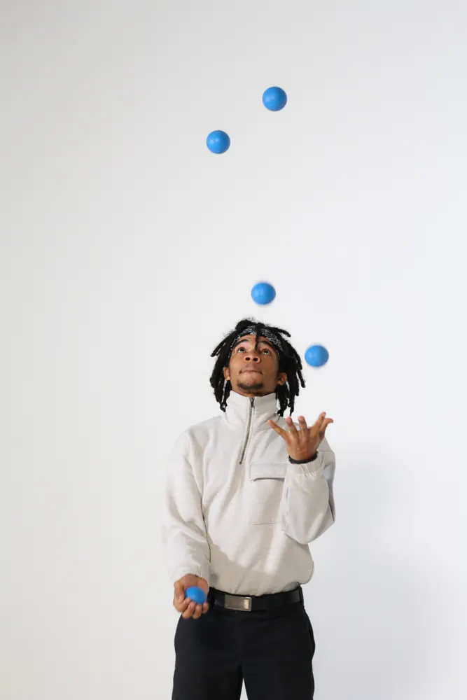 A juggler in a grey sweatshirt looking up at five blue balls in the air against a white studio wall.
