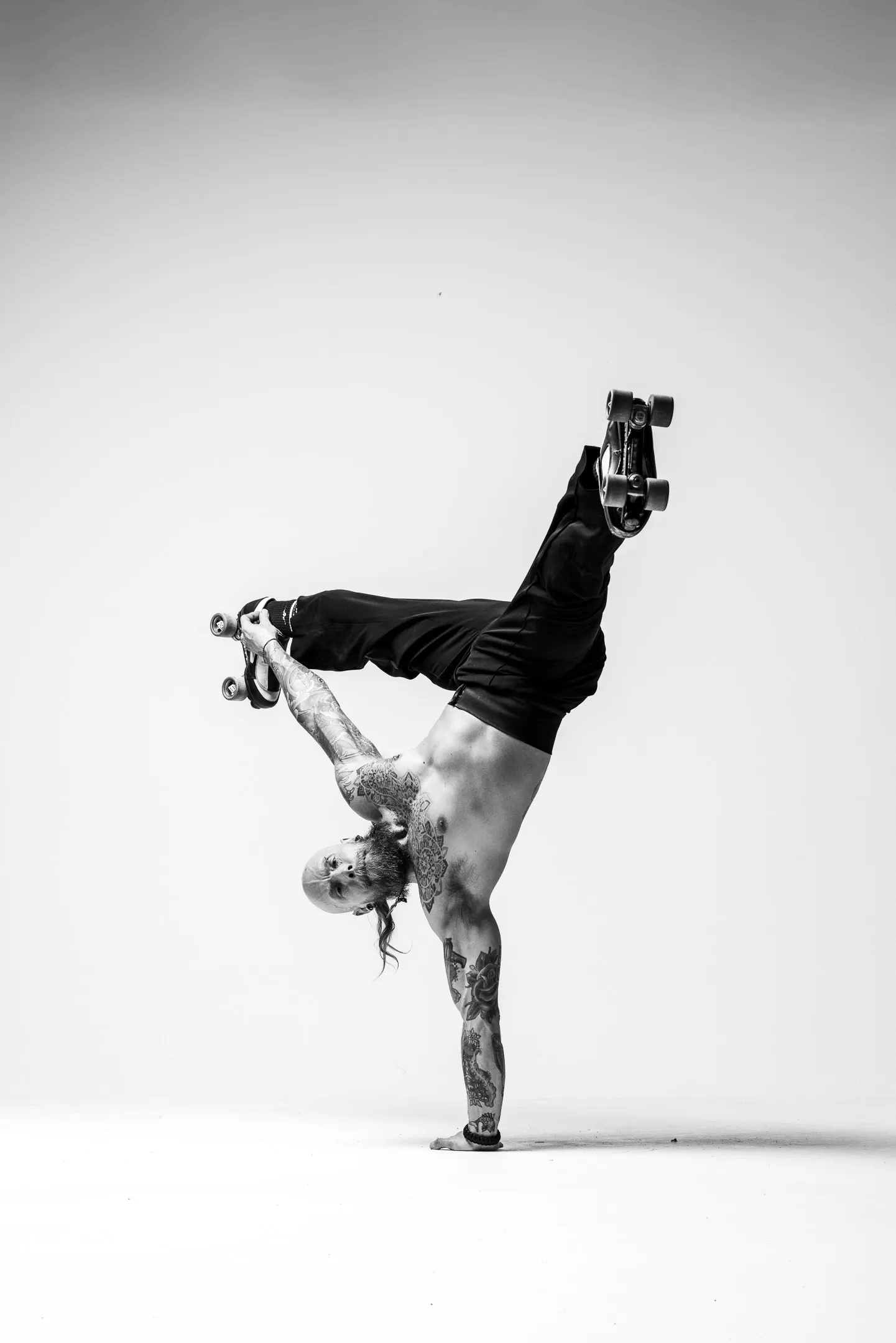 A roller skater in a one-armed handstand, legs scissored overhead with skates on, photographed in black and white in the studio.