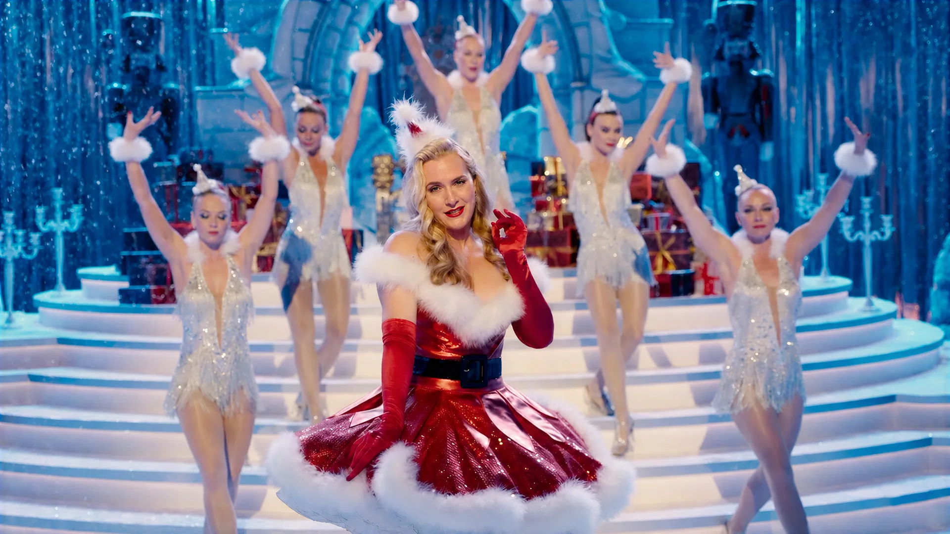 Showgirls in silver on a glittering ice-palace staircase behind a singer in a red Santa dress, in The Regime