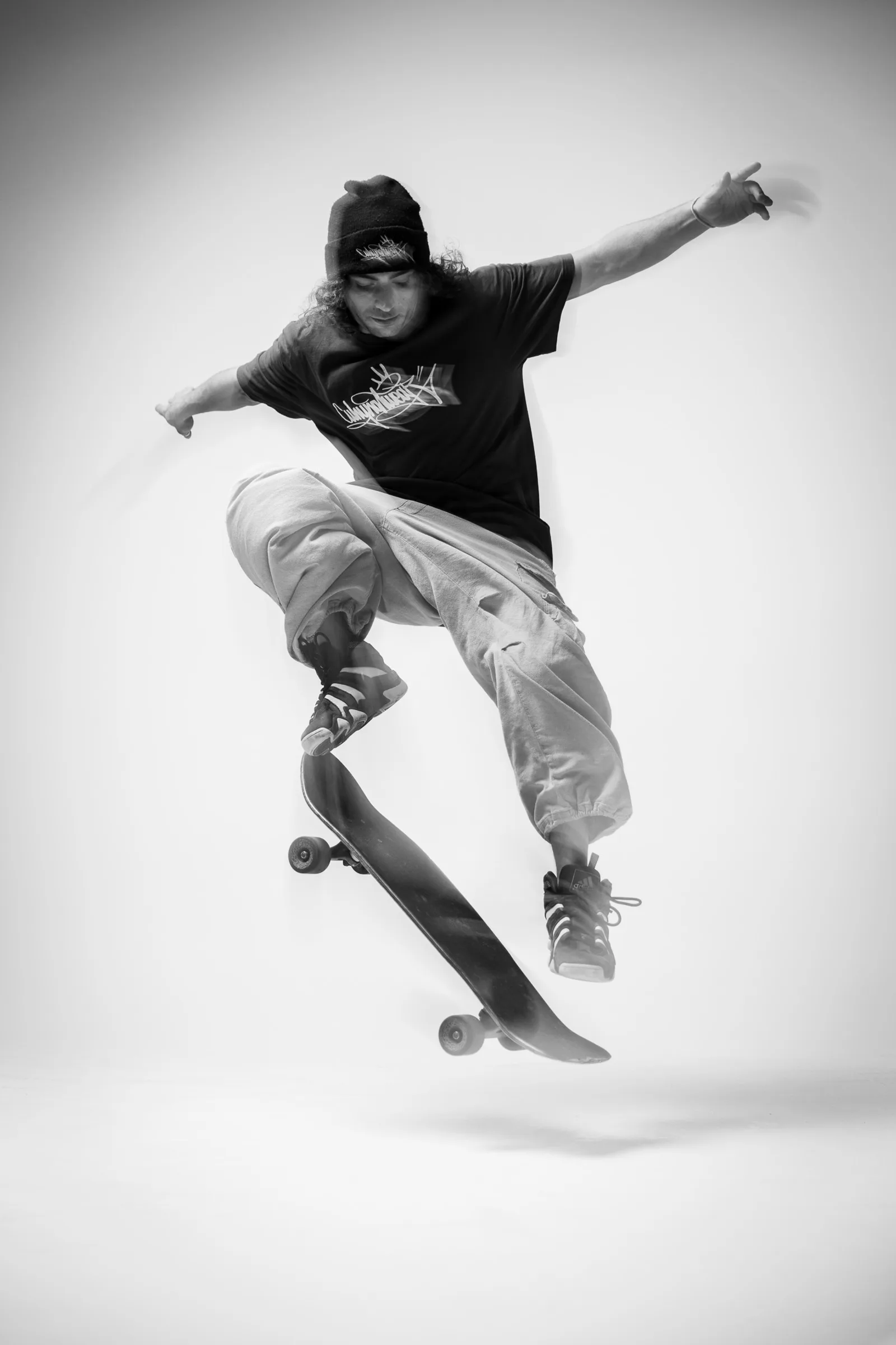 A skateboarder in a beanie and black T-shirt mid-air above his board against a white studio backdrop, arms out, the board flipping beneath him.