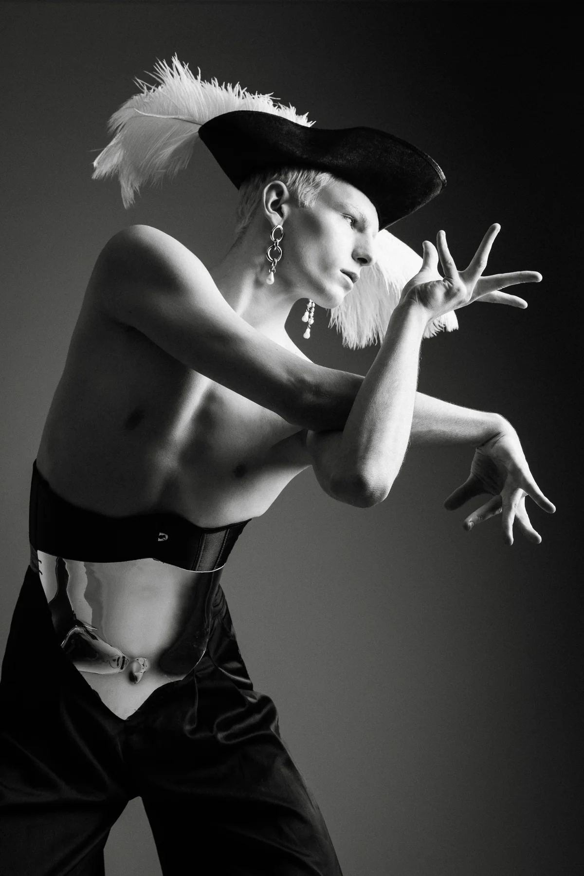 Black and white portrait of a model in a plumed tricorn hat and corset, hands raised mid-gesture