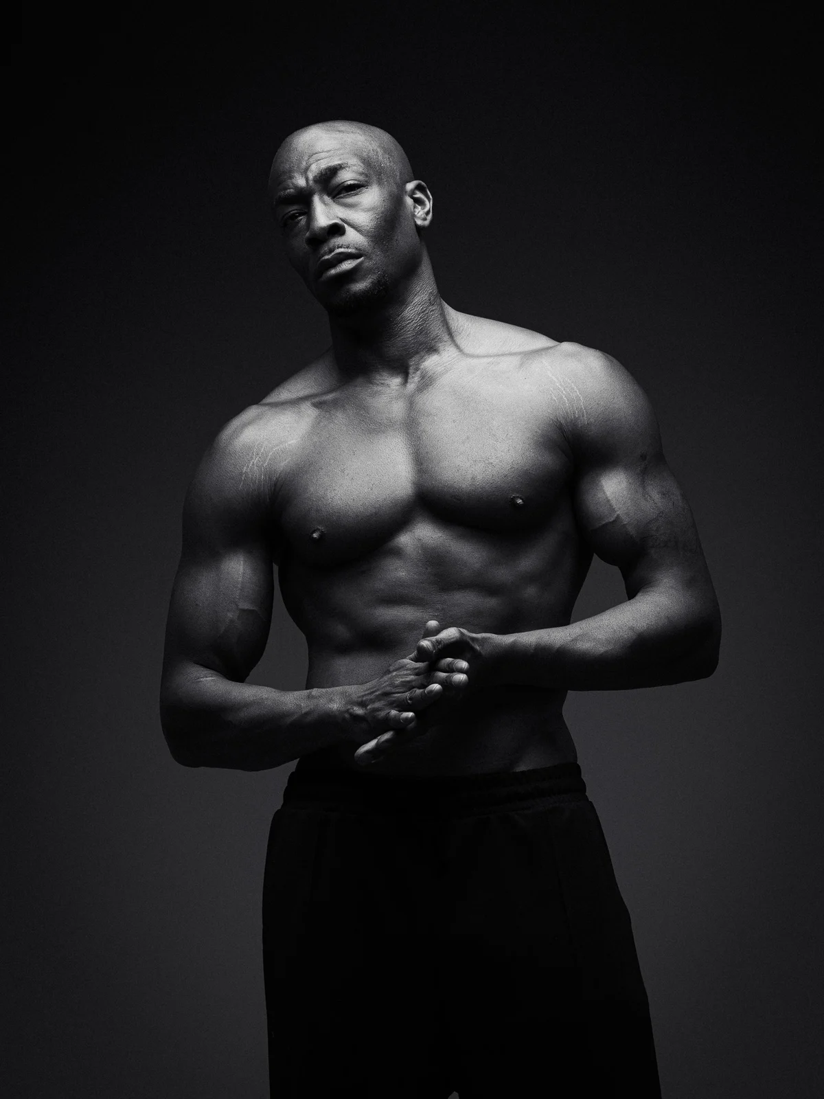 Black and white portrait of a shirtless model with a shaved head, hands clasped, against a dark studio backdrop