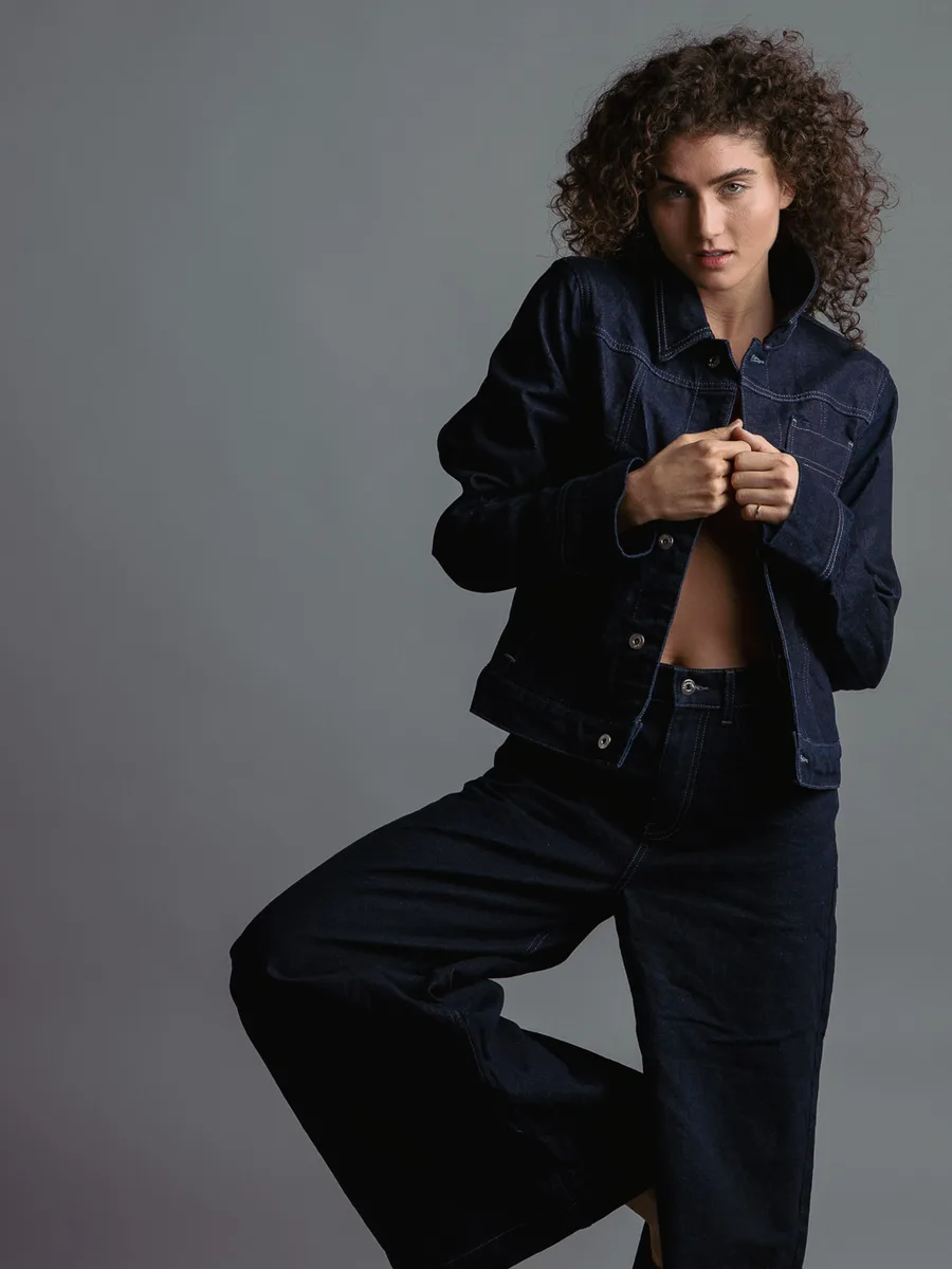 Model with curly hair in a dark denim jacket and wide jeans, standing on one leg