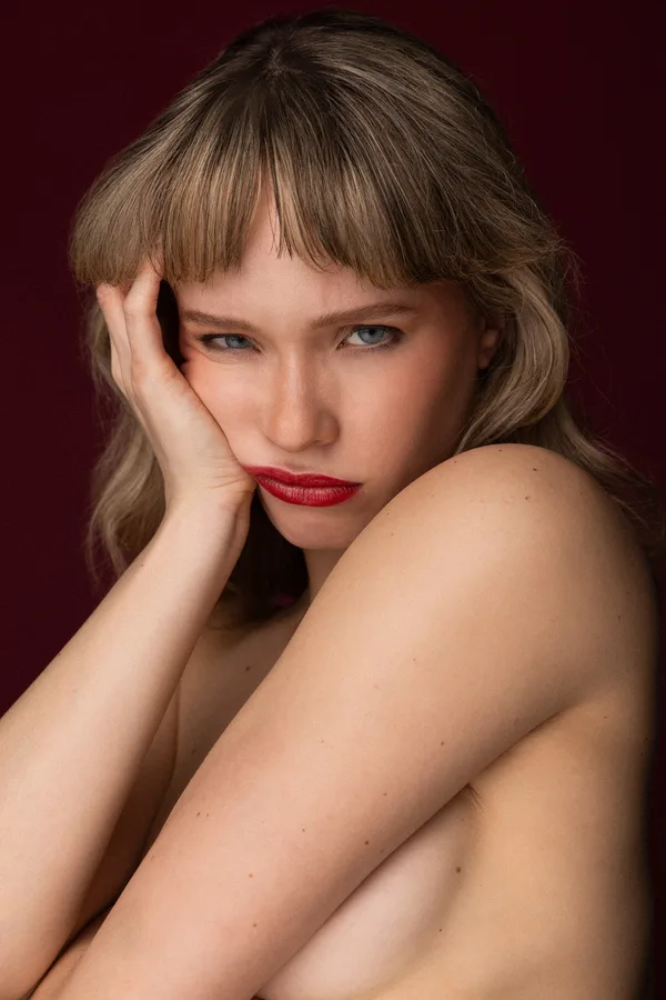 Model with a blunt fringe and red lips resting her cheek on her hand against a deep red backdrop