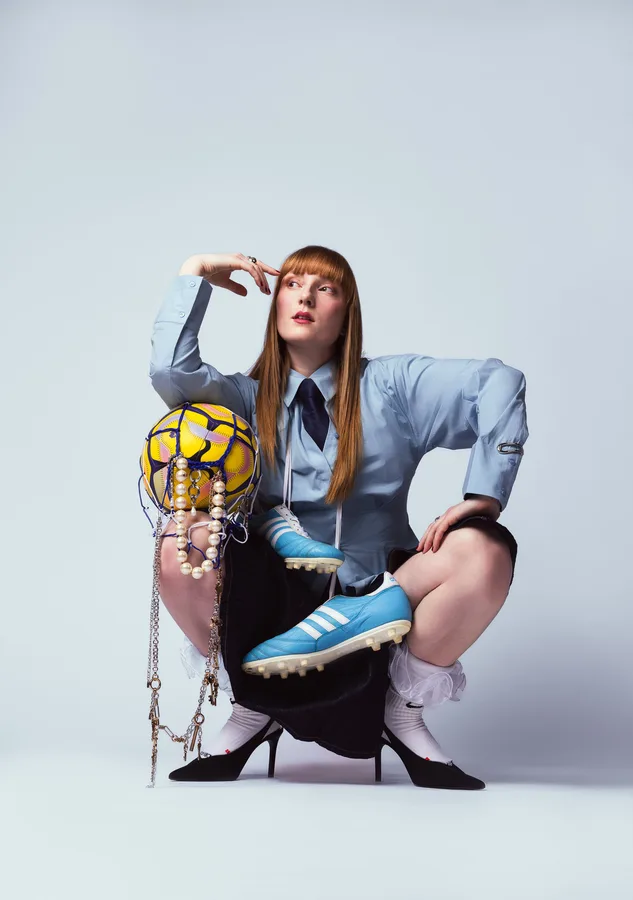 Model with long red hair crouching in a blue shirt and tie, holding a netted football