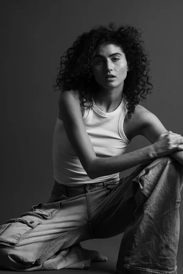 Black and white portrait of a model with curly hair in a white vest, crouching with her arms on her knee