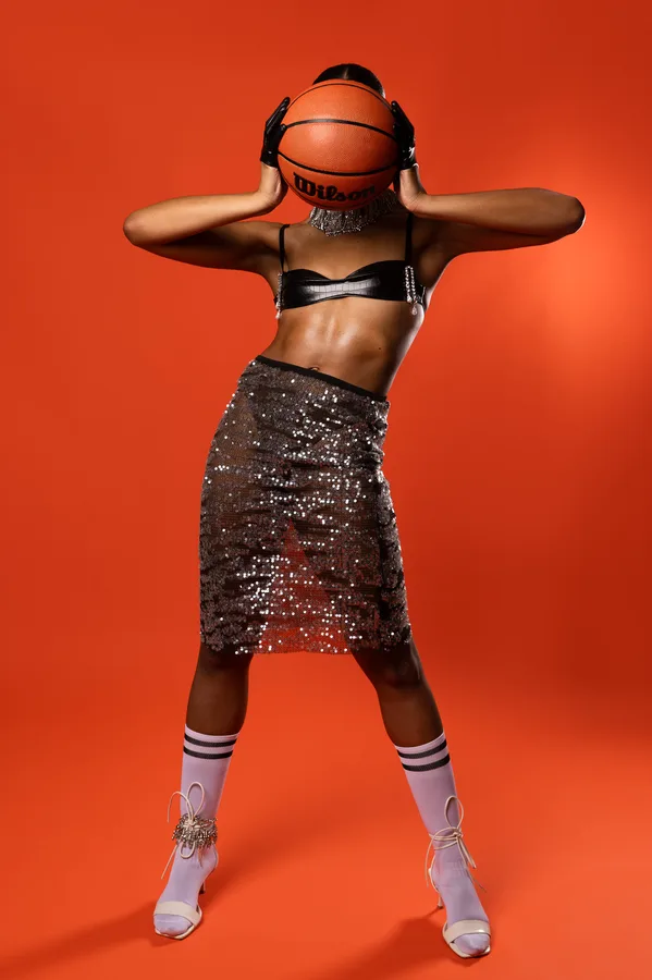 Model in a sequinned skirt holding a basketball in front of her face against an orange backdrop