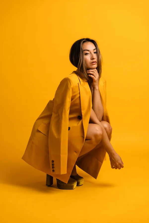 Model crouching in an oversized mustard blazer and velvet platform heels against a yellow backdrop, chin resting on her hand