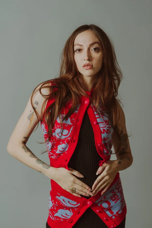 Model with long auburn hair and tattooed arms in a red patterned knitted waistcoat