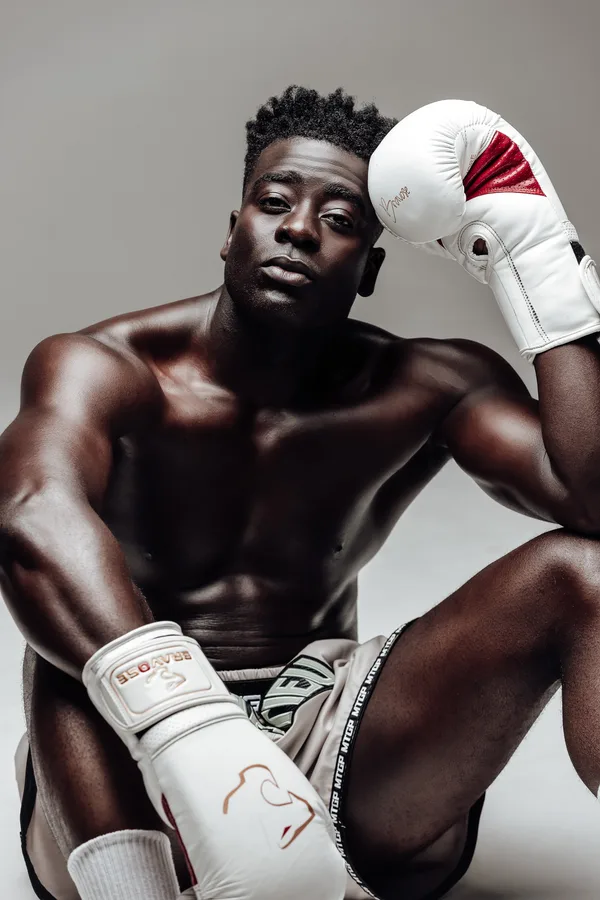 Model seated in boxing shorts, one white glove raised to his head