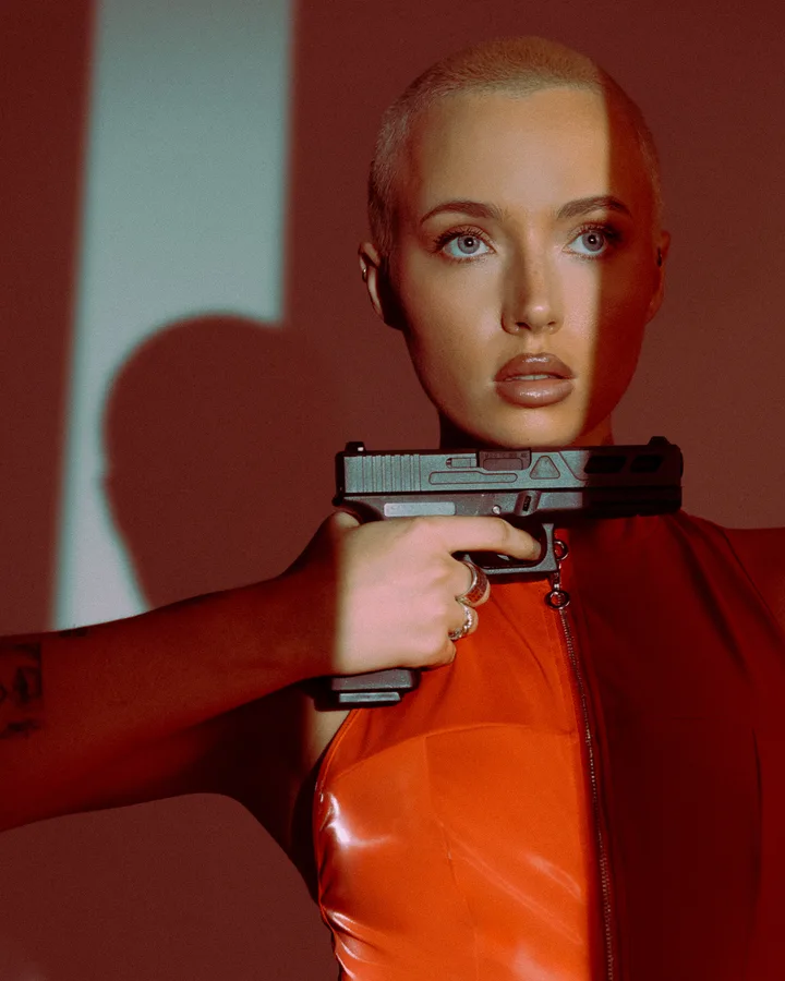 Close portrait of a model with a shaved head in a red vinyl top holding a prop pistol