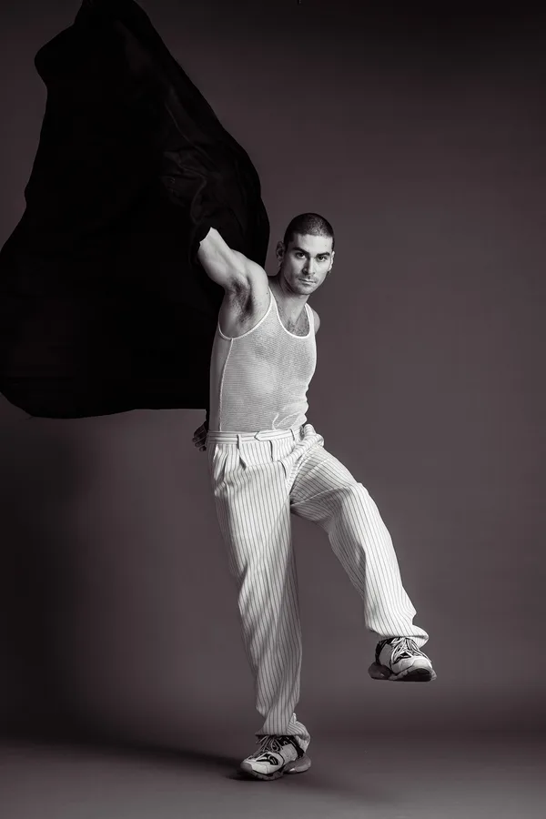Black and white shot of a model in a mesh vest and pinstripe trousers swinging a jacket behind him