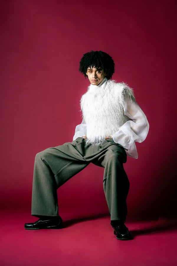 Model with curly hair in a white fringed vest and sheer sleeves, seated wide against a red backdrop