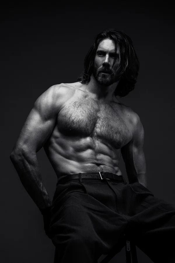 Black and white portrait of a shirtless model with long hair and a beard, seated