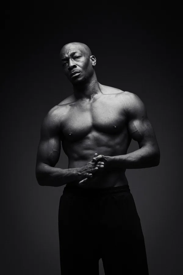 Black and white portrait of a shirtless model with a shaved head, hands clasped