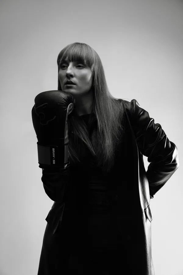 Black and white portrait of a model with a long fringe in a leather coat, a boxing glove at her chin