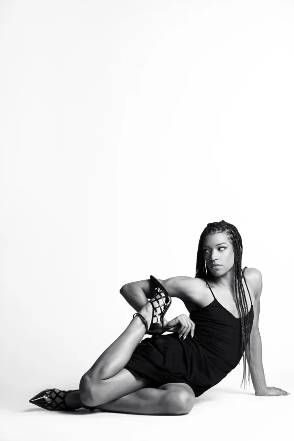 Black and white shot of a model with long braids in a black slip dress, reclining on the floor