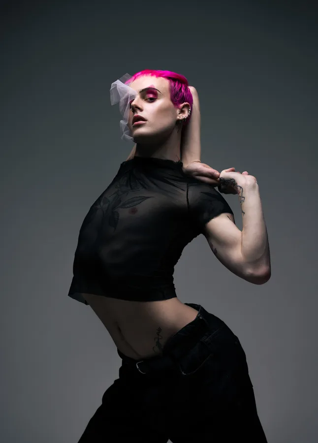 Model with pink hair and a tulle veil in a sheer crop top, one arm behind his head