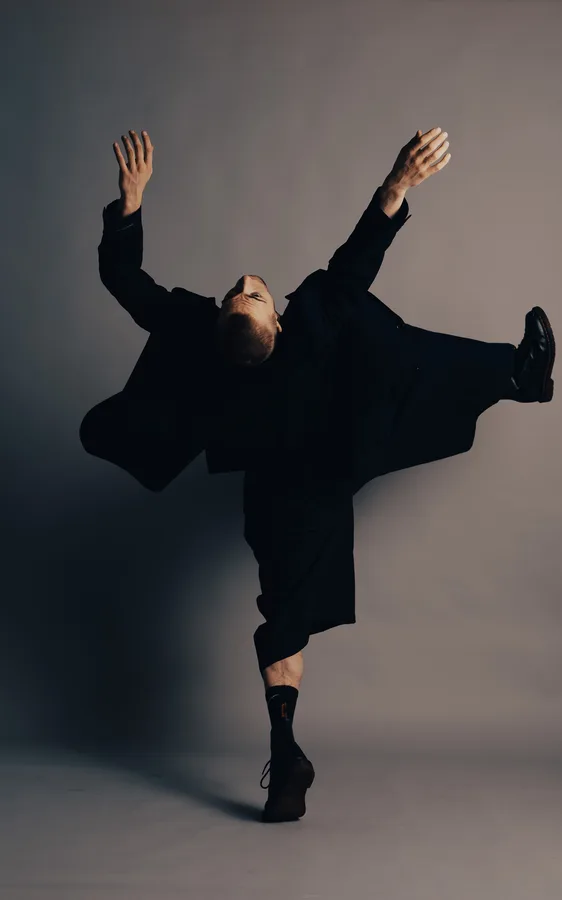 Model in an oversized black suit balancing on one leg, head thrown back and arms flung wide, against a grey studio backdrop