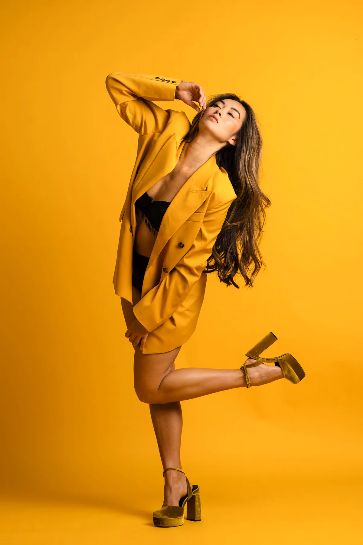 Model in an oversized mustard blazer and platform heels against a yellow backdrop, one leg kicked back