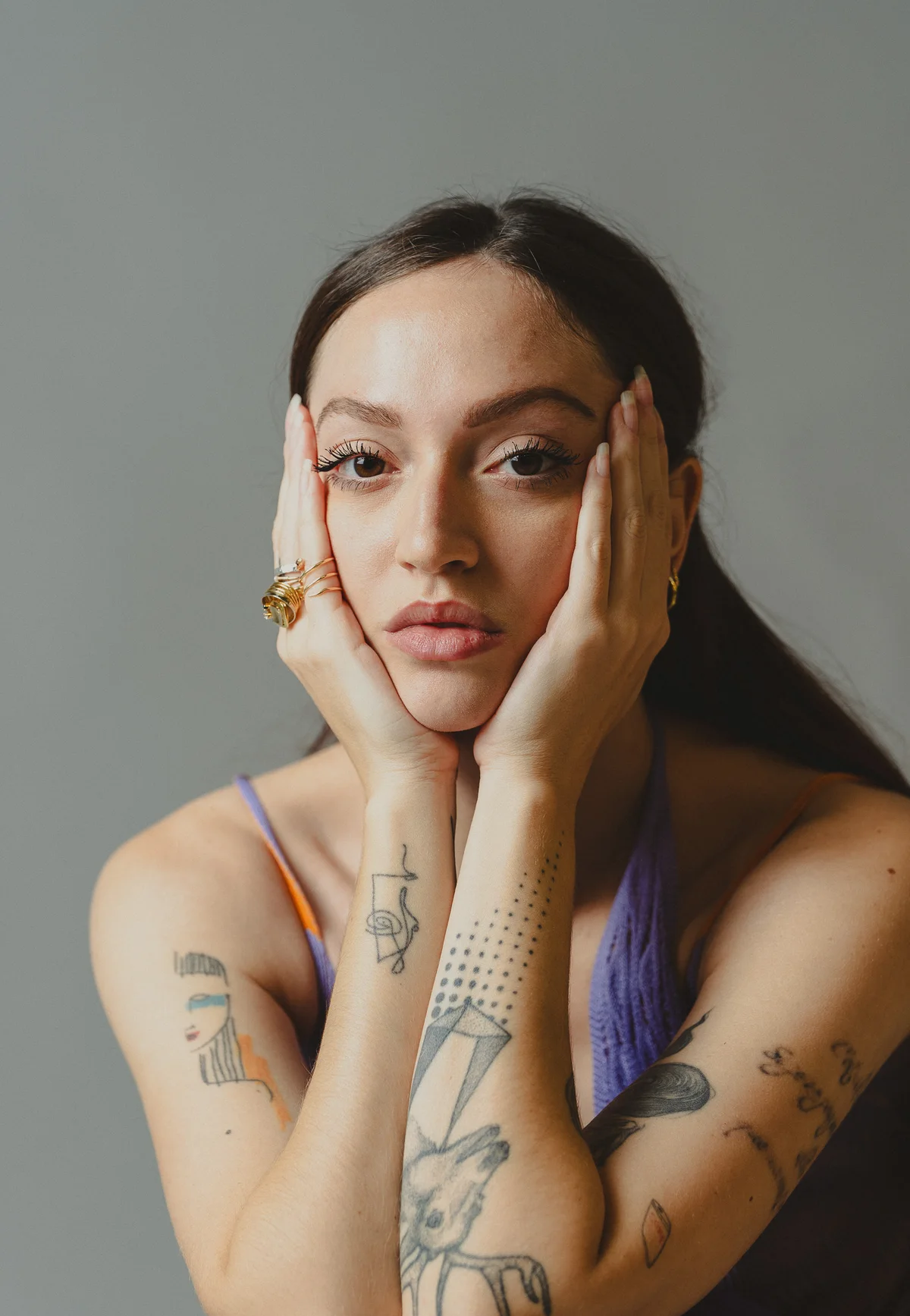 Model with tattooed arms holding her face in her hands, looking to camera