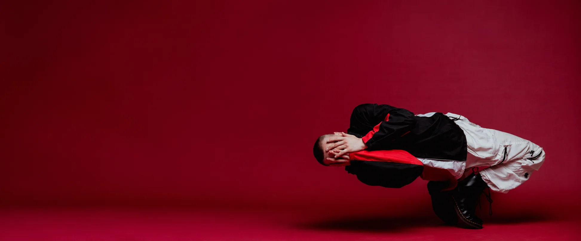 A dancer in a black and red track jacket balanced low on one hand, curled forward with his hands over his face, against a deep red studio backdrop.
