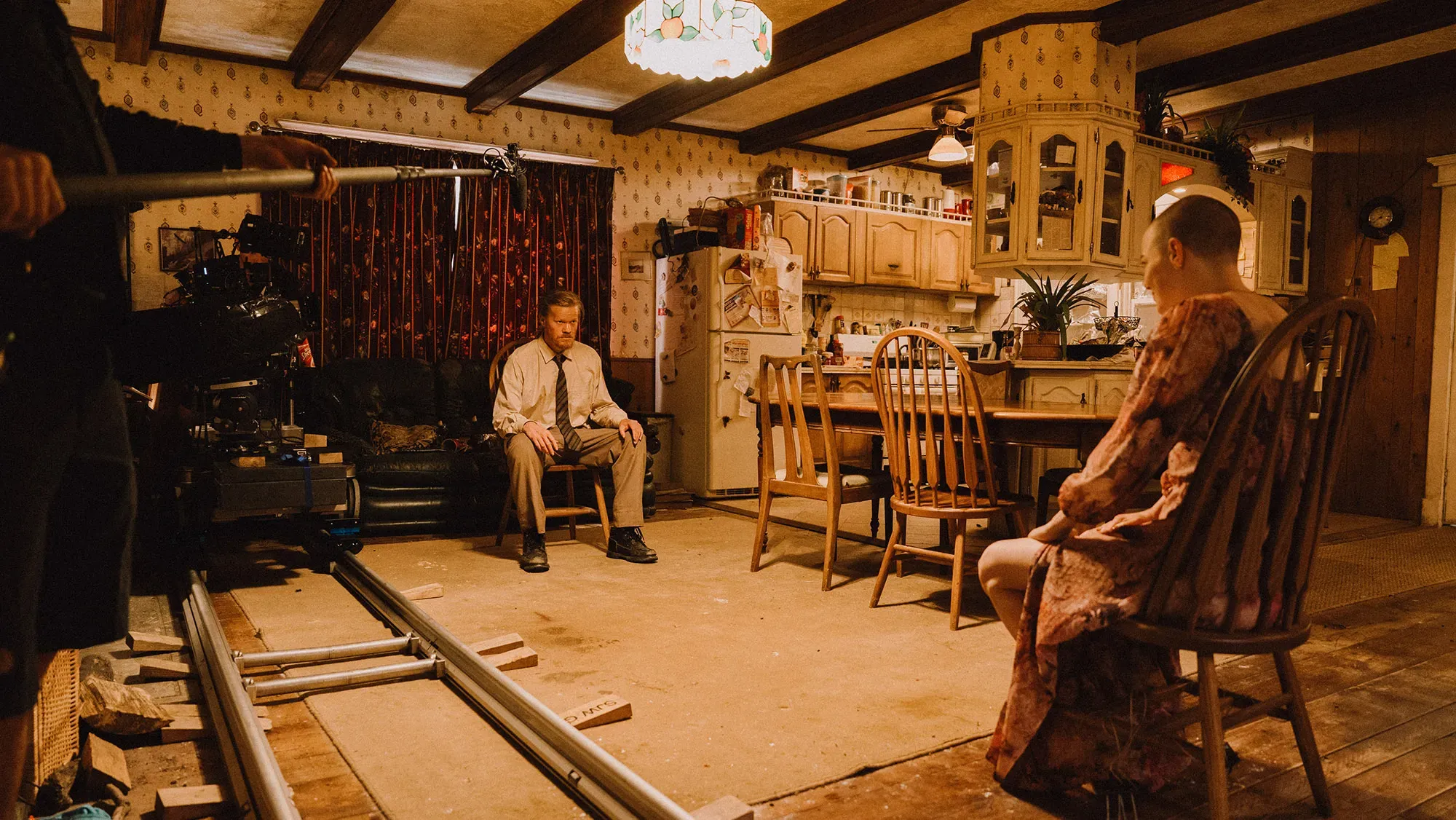 Behind the scenes on a lit kitchen set: an actor sits in shot on a wooden chair while the camera on its dolly track and a boom operator hold position