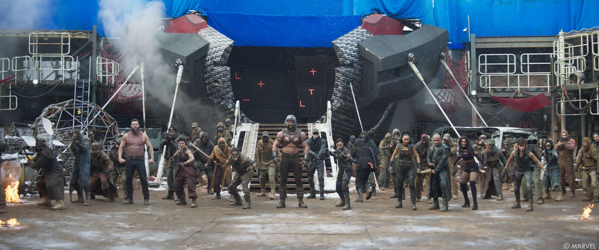 A line of costumed performers braced for action in front of a blue screen hung with tracking markers, smoke and fire on the set floor