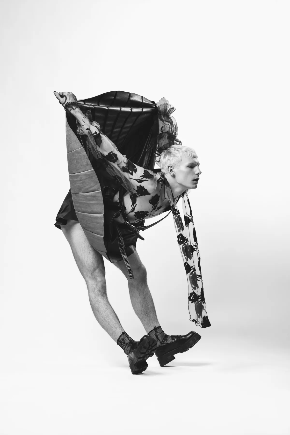 A black-and-white studio photograph of a performer folded forward at the waist, arms stretched behind them, in a sheer patterned top and heavy boots.