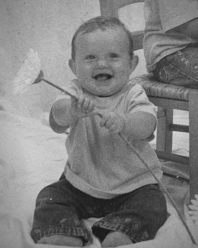 Becky as a baby, laughing and holding a daisy, sitting on a white sheet
