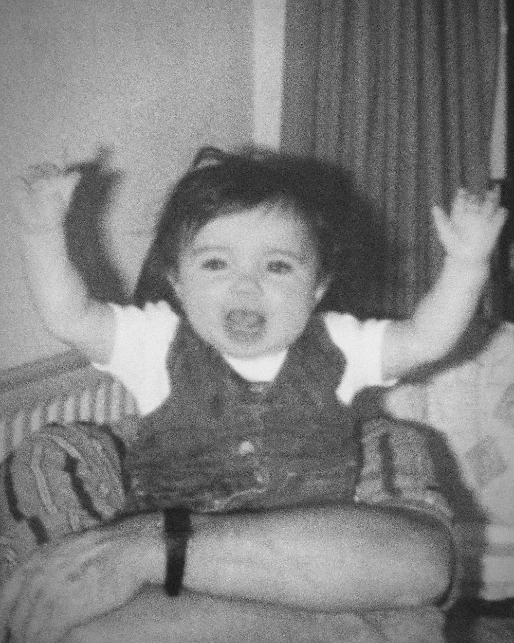 Chanel as a baby, arms in the air, sitting on someone’s lap in a denim dress