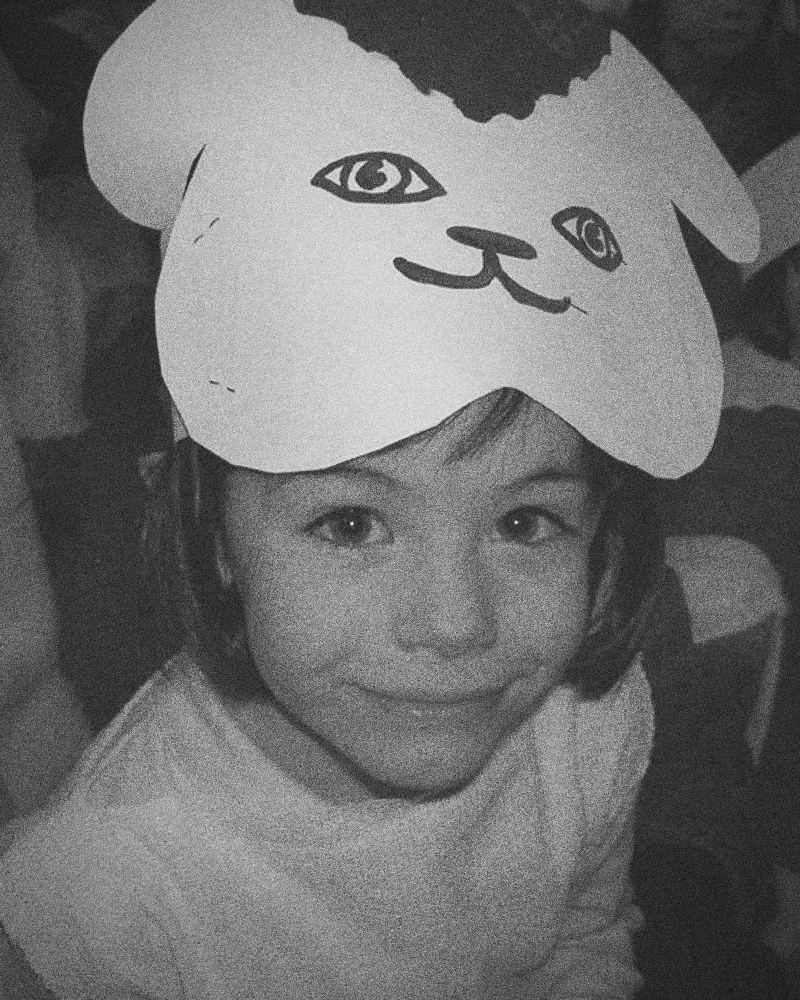 Elly as a young girl in a school play, wearing a paper animal mask on her head and smiling up at the camera