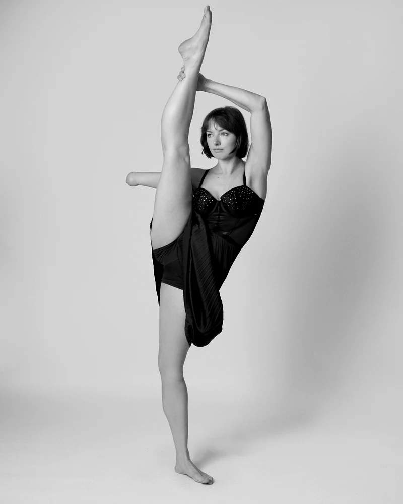 A dancer with limb difference holding one leg straight up beside her head in a studio, photographed in black and white.