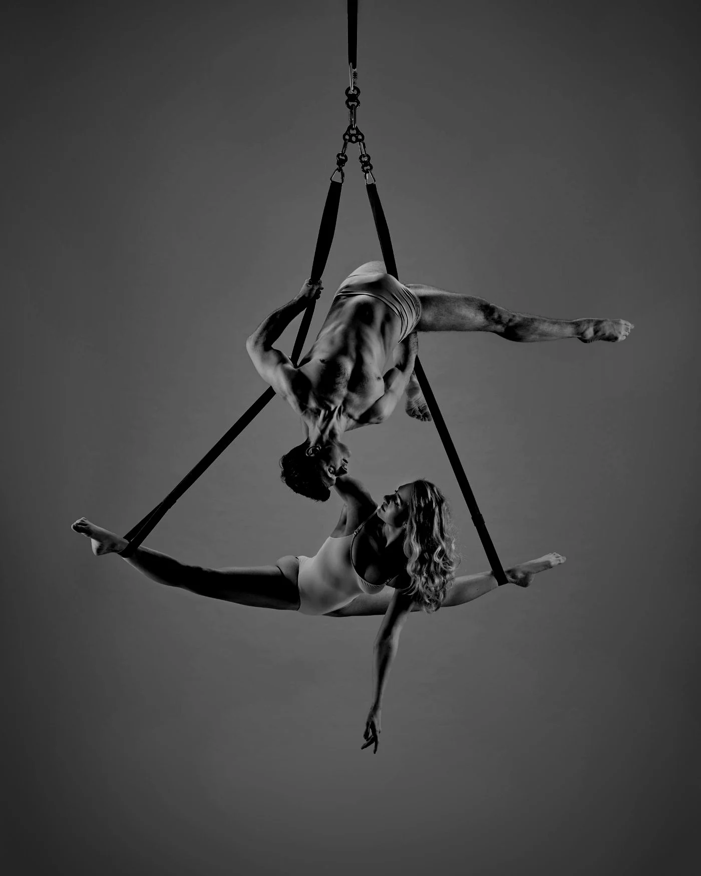 Two aerialists on straps against a dark studio backdrop, one inverted above the other in a split, their faces almost touching, photographed in black and white.