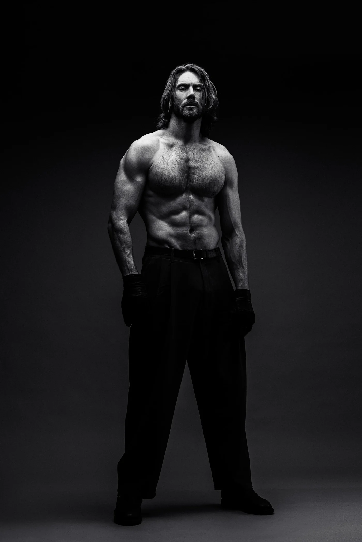 A tall, long-haired performer standing bare-chested in dark trousers and gloves, lit from above against a dark studio backdrop, photographed in black and white.