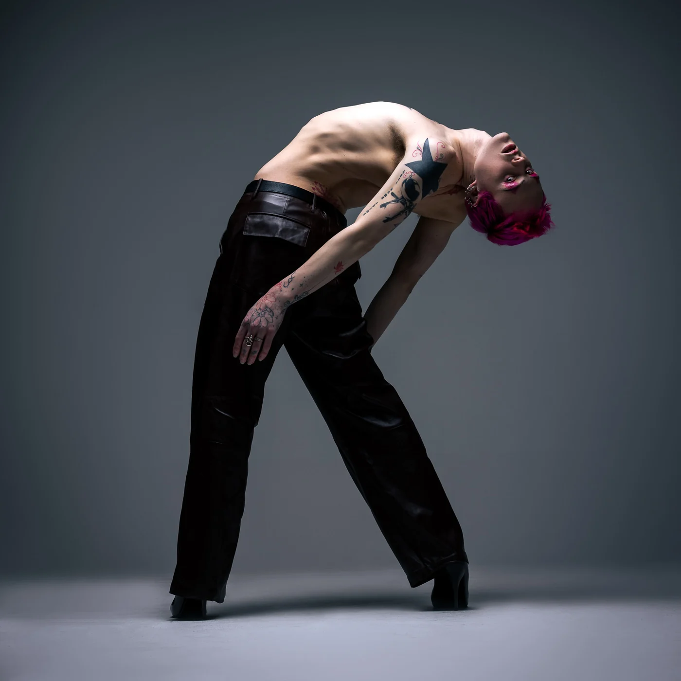 A shirtless performer with pink hair and tattooed arms in leather trousers and heeled boots, bent into a deep backbend with arms hanging loose, against a grey studio backdrop.