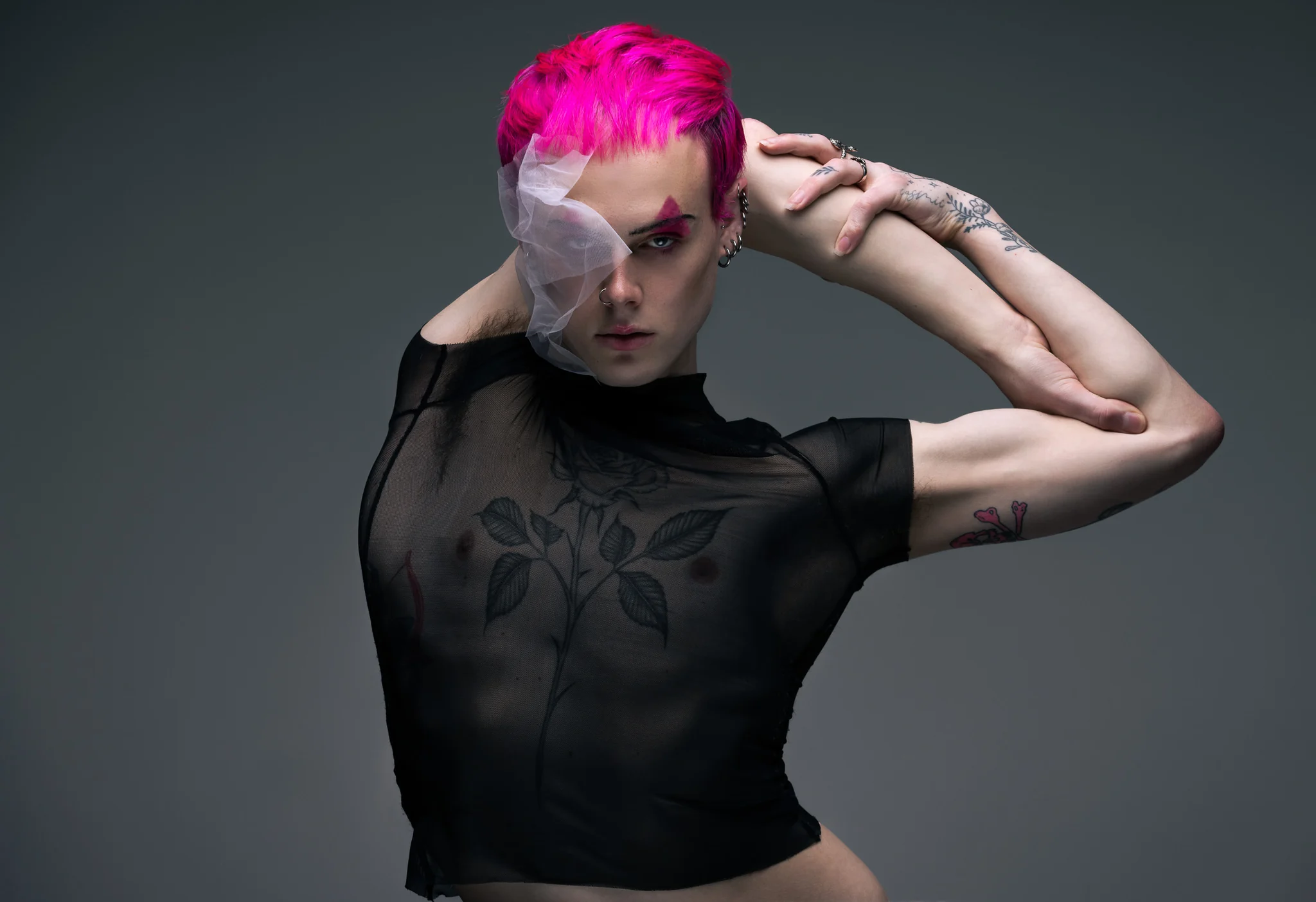 A performer with bright pink hair and a veil of white tulle over one eye, in a sheer black top over a rose tattoo, one arm folded behind the head, against a grey studio backdrop.