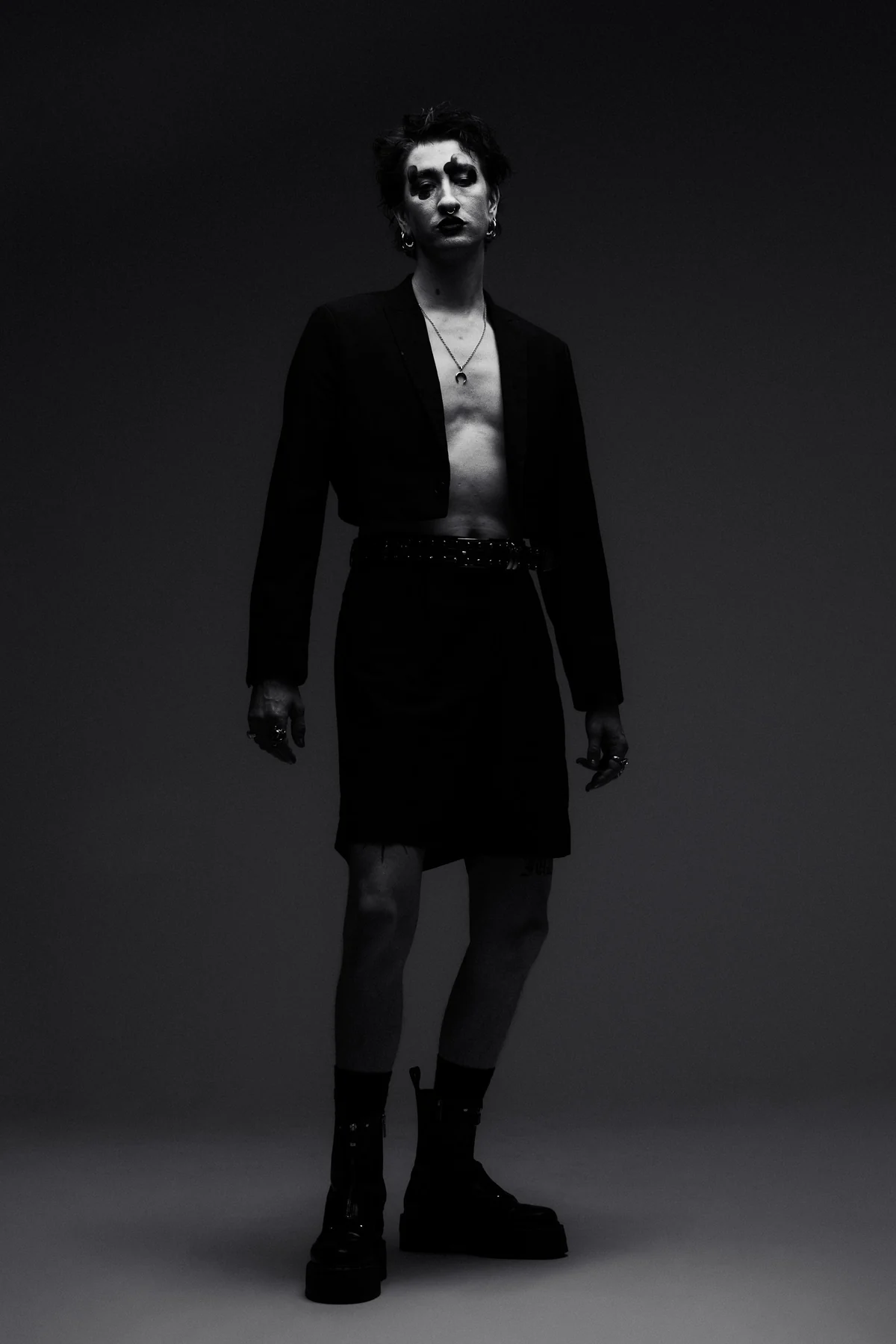 A full-length studio portrait of a performer with painted eyes, rings and a pierced lip, in a cropped jacket, skirt and platform boots, photographed in black and white against a dark backdrop.